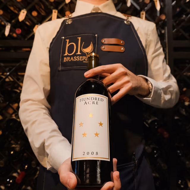 A sommelier in a Blu Brasserie apron holding a bottle of Hundred Acre 2008